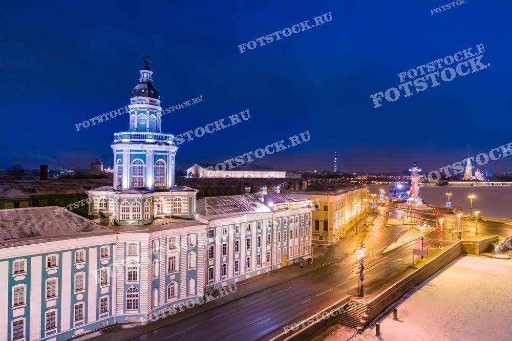 На фотографии изображен вечерний зимний вид на Кунсткамеру в Санкт-Петербурге, подсвеченную и выделяющуюся на фоне ночного города. Освещенные улицы и отражение городских огней в реке Неве добавляют изображению ощущение тепла и спокойствия, несмотря на зимний холод.